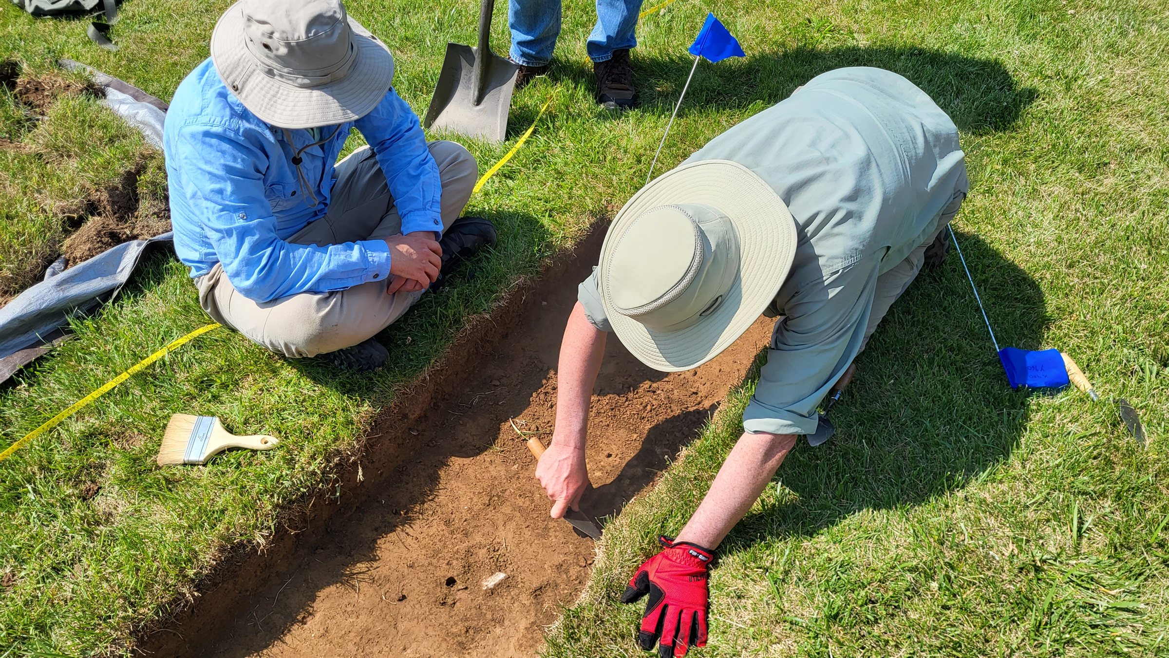 Two researchers ground-truthing a geophysical anomaly in a small test unit at Old Georgetown Cemetery, with pin flags, trowel and brush